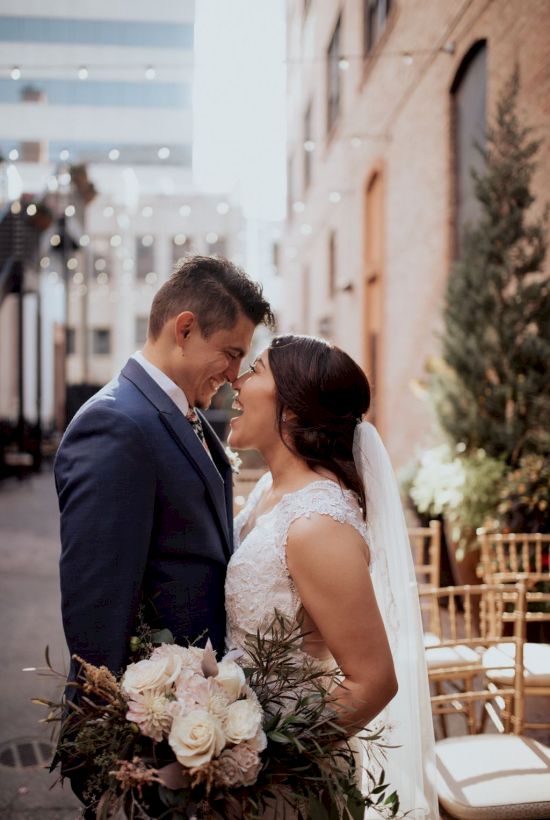 The image shows a bride and groom standing close together outdoors, facing each other. The bride holds a bouquet, and both are smiling.