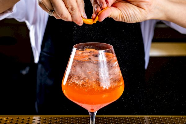 A person is preparing a cocktail, holding an orange twist over a glass with an orange beverage, smiling, using marble and brass barware.