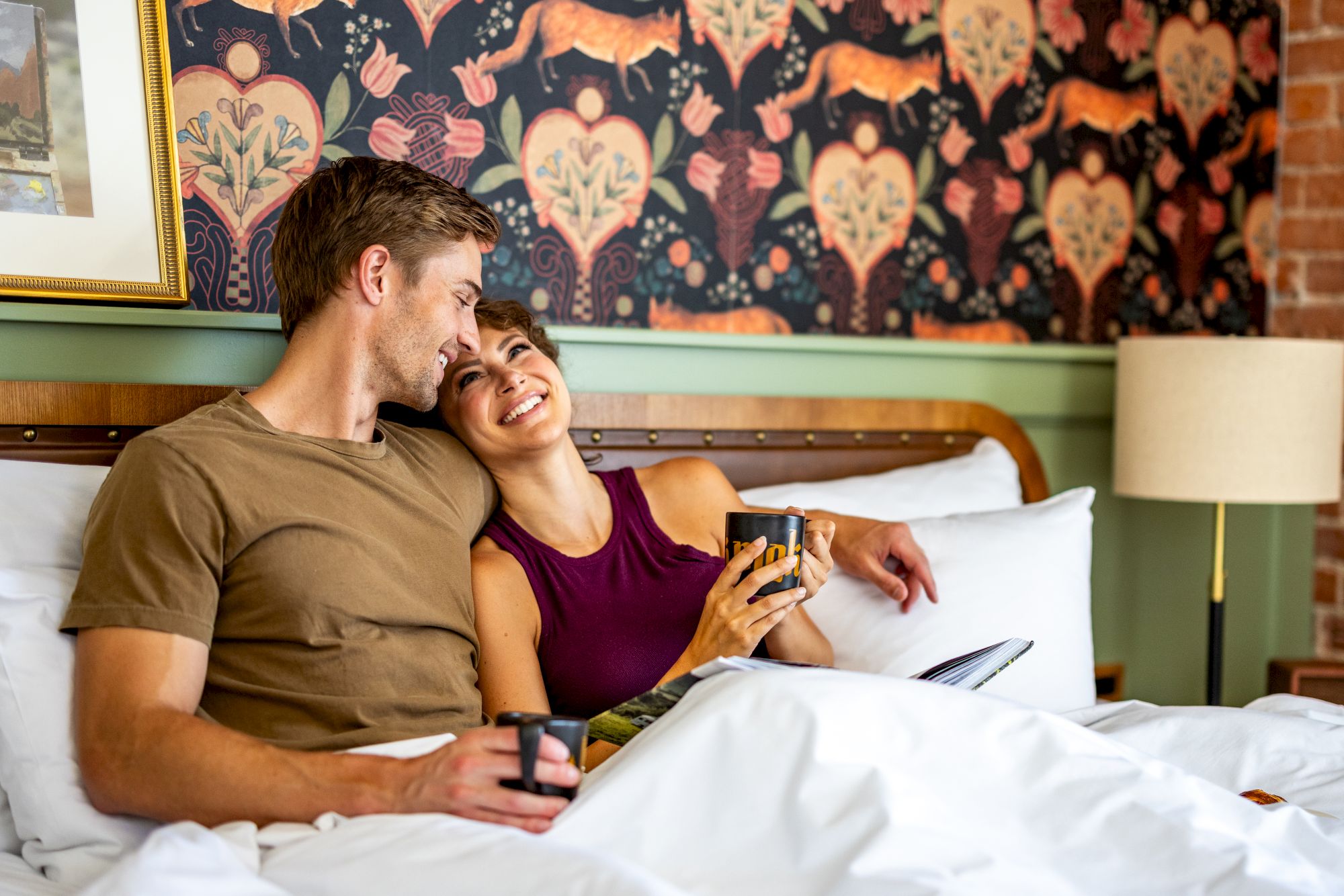 Two people are sitting in bed, smiling and holding coffee mugs, in a cozy room with patterned wallpaper and soft lighting.