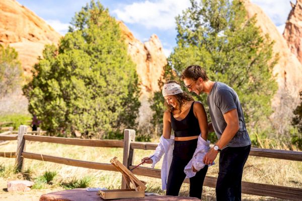A couple is enjoying an outdoor activity with a wooden game, set against a scenic background of trees and rocky formations.