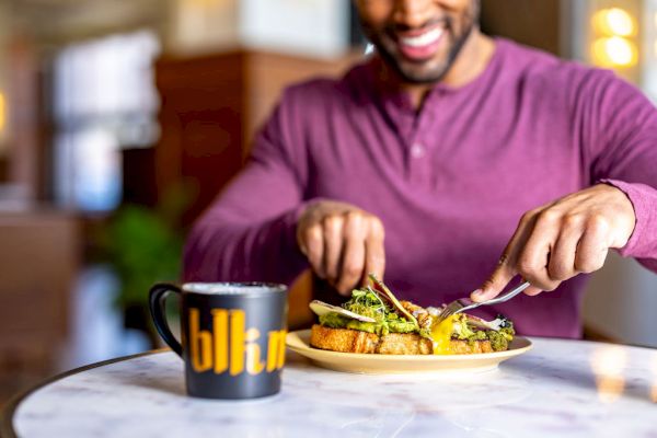 A person in a purple shirt is smiling while cutting food on a plate, with a cup labeled 