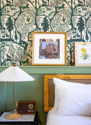 A cozy bedroom corner with patterned wallpaper, framed art, a bed with white pillows, a bedside table with a retro lamp and a vintage radio.