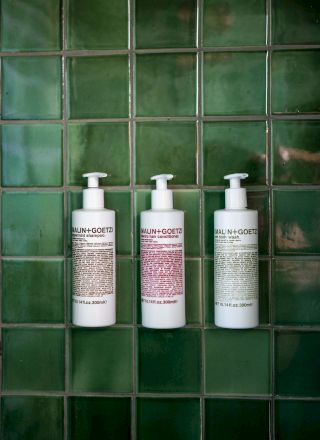 Three white pump bottles with text are mounted on a dark green tiled wall. They appear to be toiletries, likely shampoo, conditioner, and body wash.