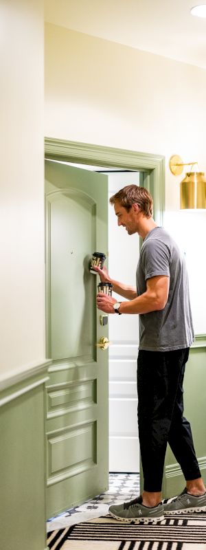 A person is opening a door in a hallway with light green walls, patterned carpet, and gold lighting fixtures above the doors.