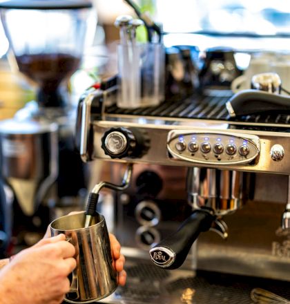 A person is using a coffee machine, frothing milk with a steam wand, likely in a cafe or coffee shop setting.