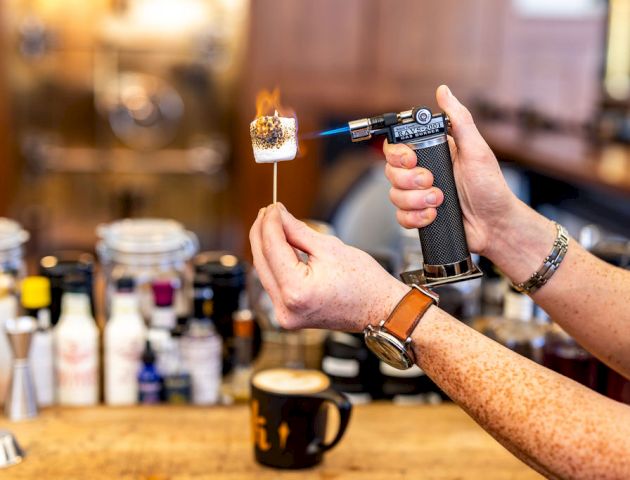 A person is using a torch to roast a marshmallow on a stick, with various items and a cup of coffee on the counter.