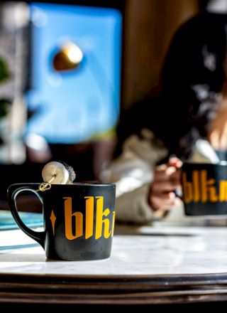 A person holding a mug sits at a table with another mug and a book in sunlight.