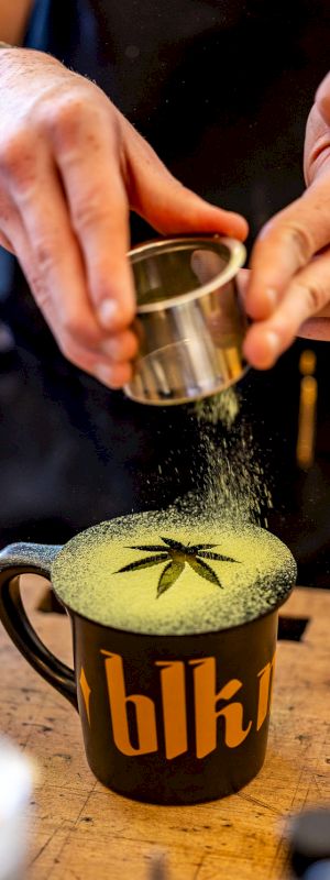 A person dusting green powder, likely matcha, over a mug with a leaf design on top. The mug has the letters 