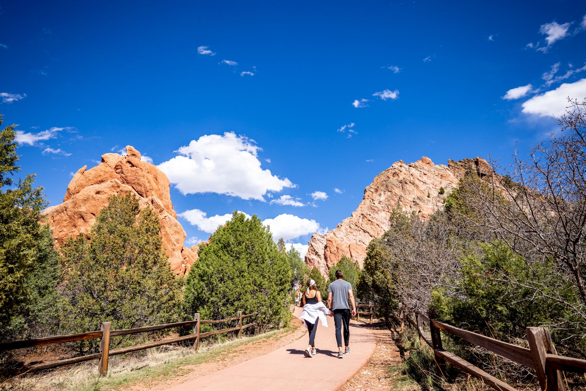 A couple walks down a scenic trail flanked by trees and red rock formations under a blue sky with scattered clouds, enjoying a sunny day outdoors.