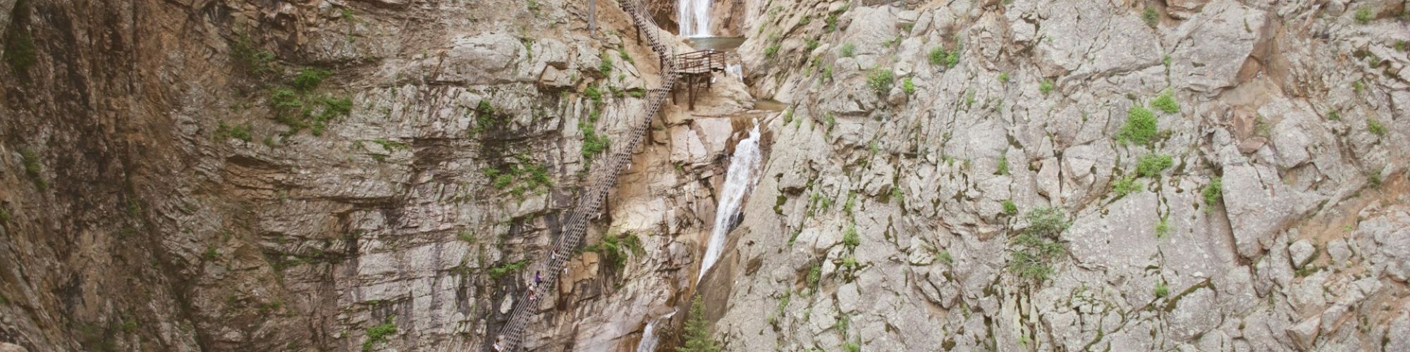 The image shows a waterfall cascading down a rocky cliff into a small pool, with people visible at the base. Pine trees dot the area.
