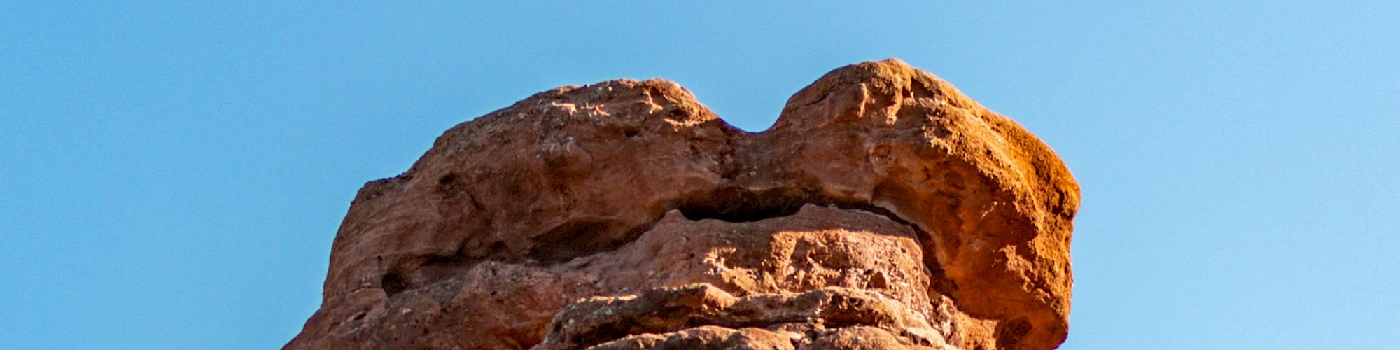 A tall, red rock formation under a clear blue sky, with layers of rock visible, likely part of a larger geological landscape.
