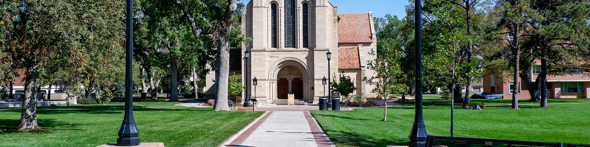 A pathway leads to a church with an arched entrance surrounded by trees and lampposts, with benches on either side and a clear blue sky.
