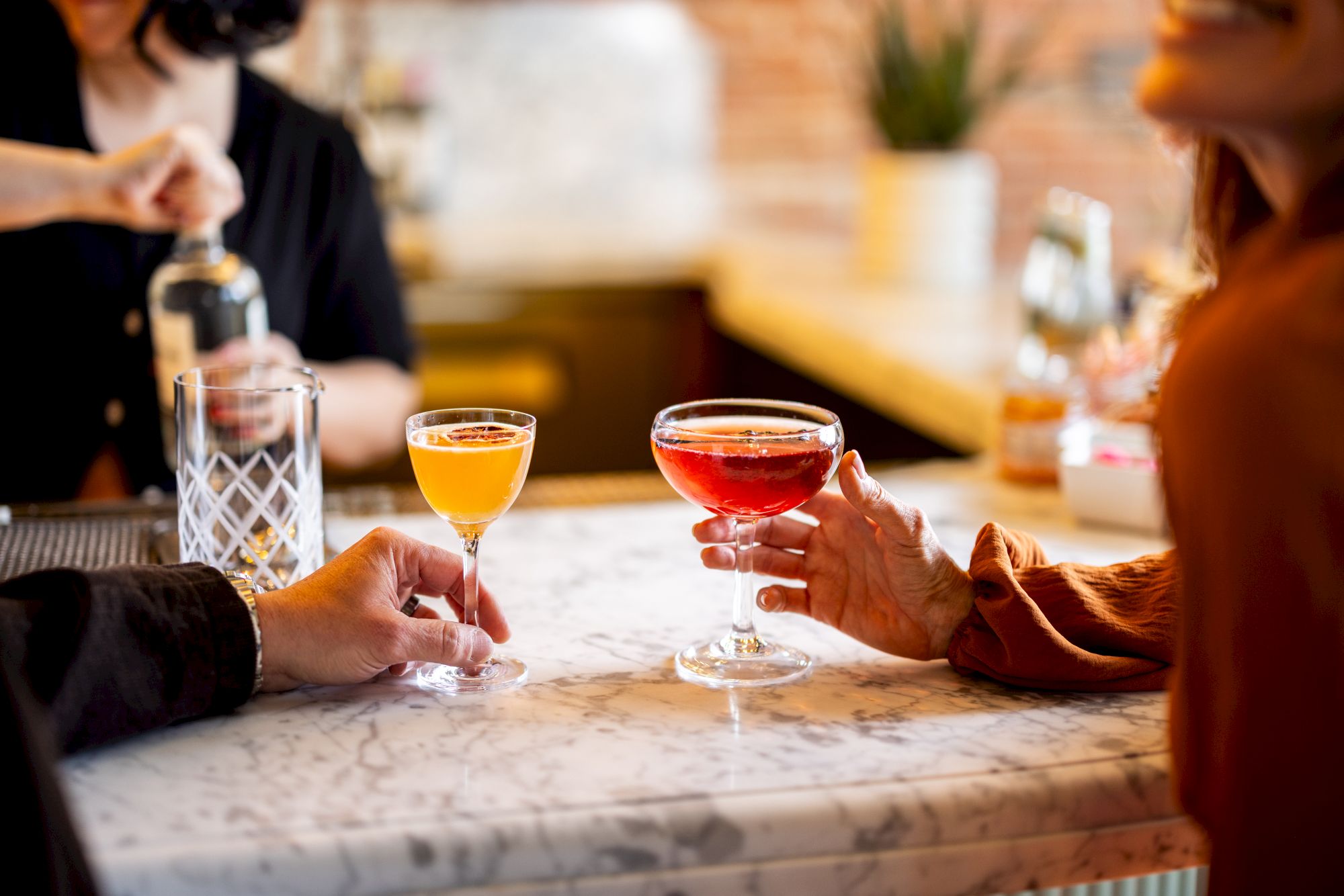 Two people are sitting at a marble bar, each holding a cocktail. One drink is yellow, and the other is red. A bartender is in the background.