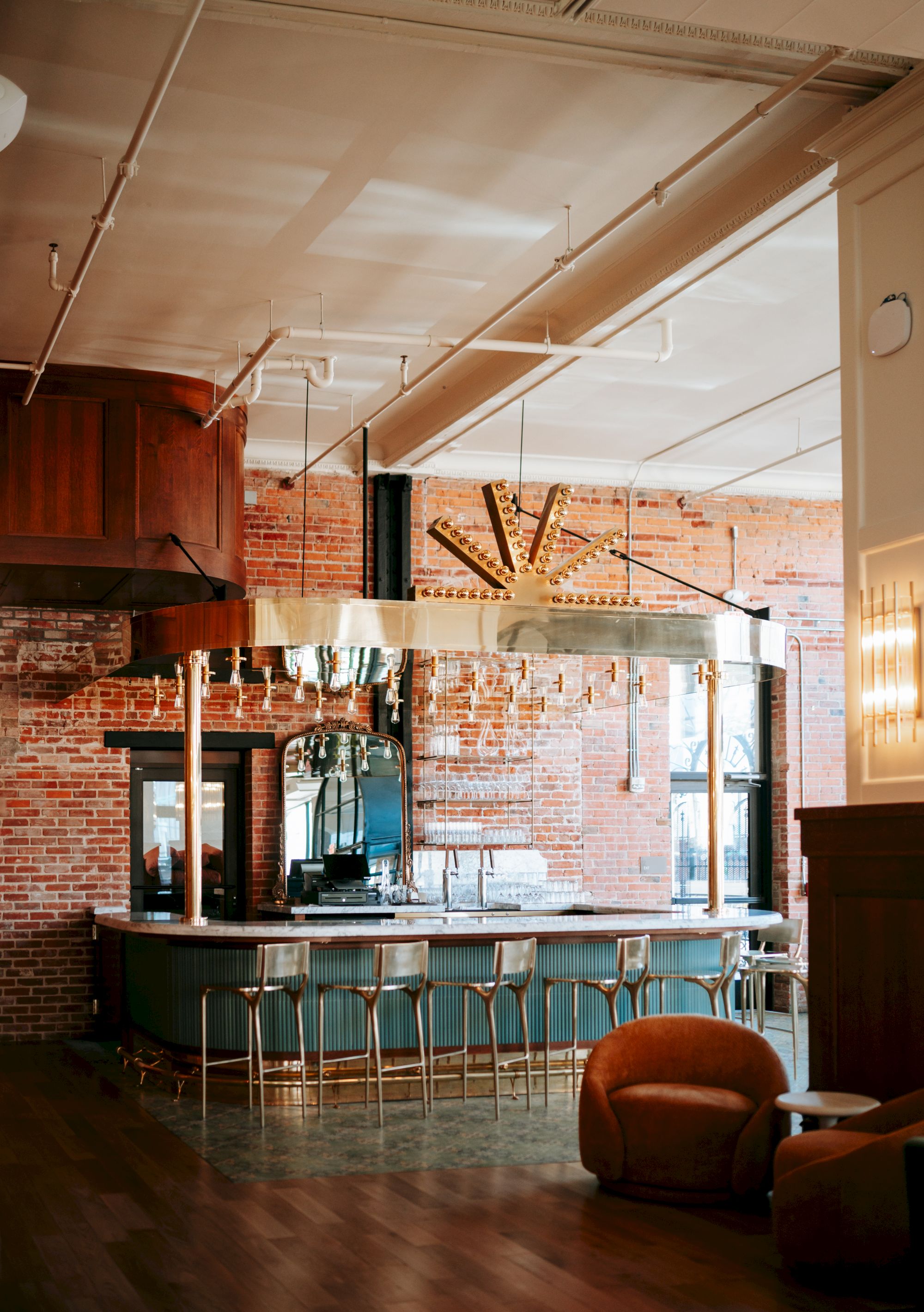 A modern bar with brick walls, a wooden ceiling, bar stools, a mirrored backdrop, and a cozy seating area with a plush armchair is shown.