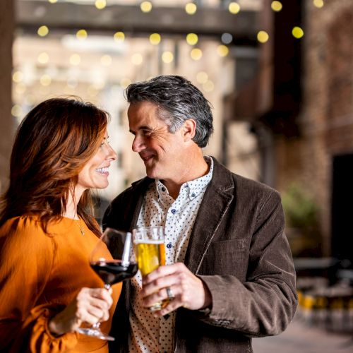 A couple is standing close together in an outdoor setting, each holding a drink and smiling at each other with string lights in the background.