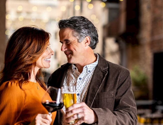 A couple is standing close together in an outdoor setting, each holding a drink and smiling at each other with string lights in the background.