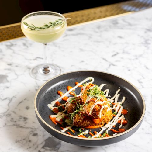 A cocktail garnished with a sprig of rosemary is beside a dish of food with sauce drizzle, herbs, and colorful garnish on a marble table.