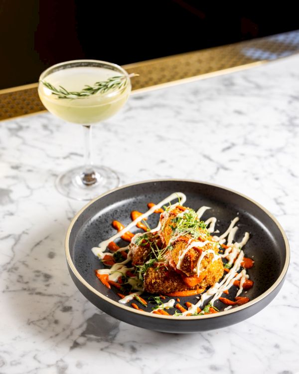 A cocktail garnished with a sprig of rosemary is beside a dish of food with sauce drizzle, herbs, and colorful garnish on a marble table.