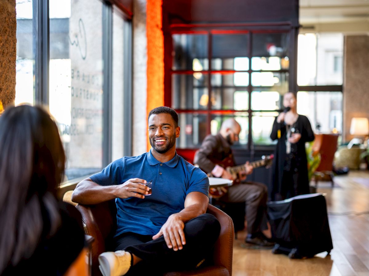 A man is sitting on a chair, smiling and holding a drink, while musicians perform in the background in a modern indoor setting.