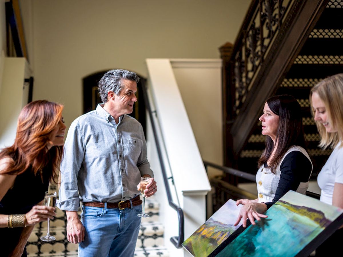 A group of people engages in conversation while viewing artwork inside a building with a stairway in the background, possibly at an art exhibit.