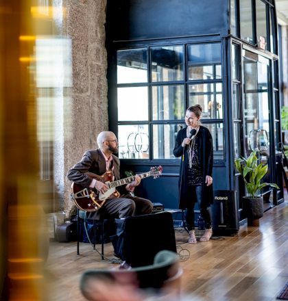 A musician plays the guitar while a singer performs beside him in a stylish indoor setting with large windows and modern decor.