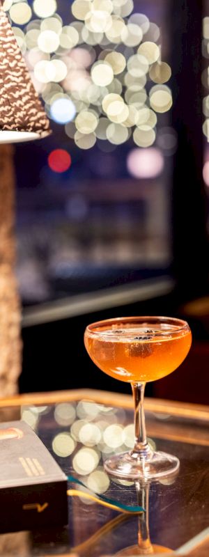 A cocktail sits on a glass table next to a book and a lamp, with bokeh lights in the background.