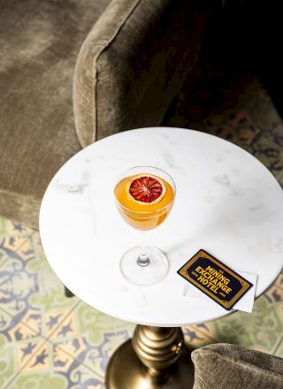 A round marble table with a cocktail garnished with an orange slice, a glass, and a black card with gold text, next to a patterned floor and a sofa.