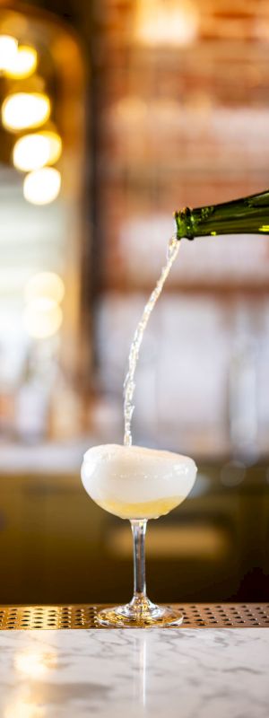 A bottle is pouring a drink, likely champagne or sparkling wine, into a coupe glass on a bar counter with a blurred background of lights.