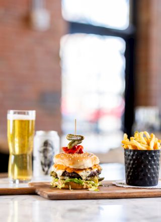 A burger on a wooden board, fries in a metal container, a glass of beer, and a beer can on a marble counter with a blurred background.