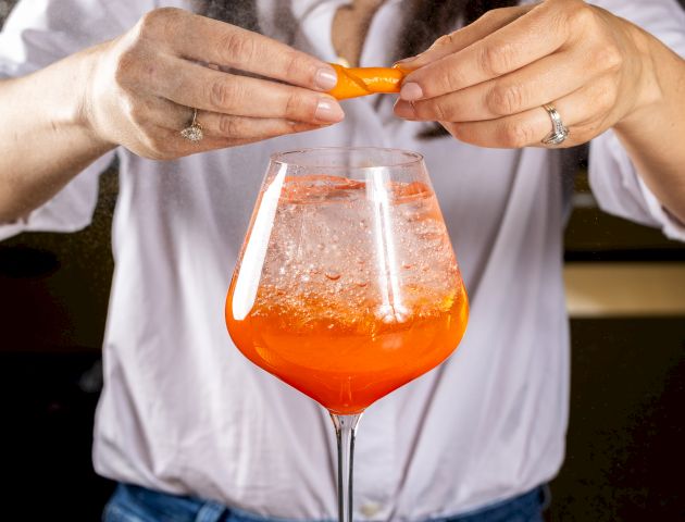 A person is squeezing an orange peel over a cocktail in a large wine glass, likely adding a touch of zest.