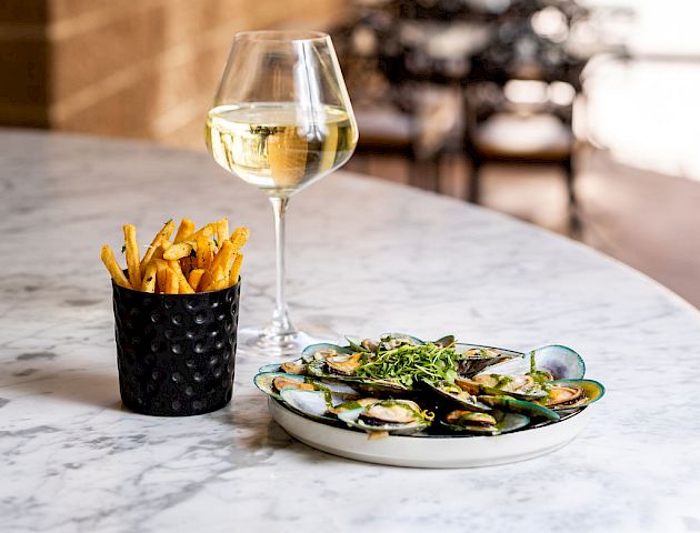 A table set with a glass of white wine, a small black cup of fries, and a plate of green mussels. The background shows outdoor seating and a building.