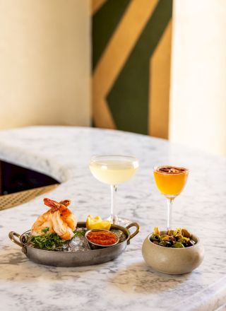 The image shows two cocktails in elegant glasses and a plated seafood dish with garnishes on a marble table, alongside a bowl of greens.