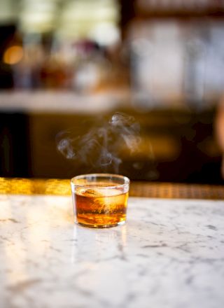 A glass of amber-colored liquid on a marble surface, with smoke rising from it, set against a blurred background indoors.