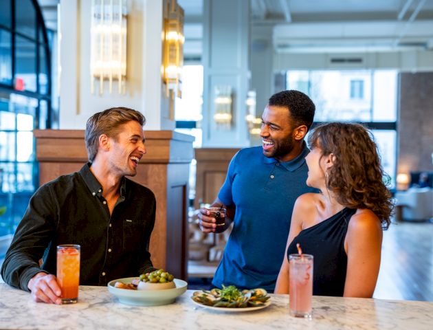 Three people are enjoying drinks and appetizers at a well-lit, stylish indoor setting, engaging in lively conversation.