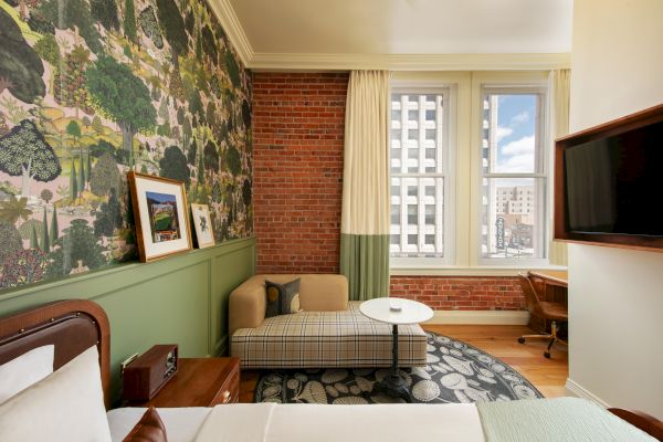 The image shows a cozy room with a bed, a patterned wallpaper, exposed brick wall, sofa, round table, TV, desk, and large windows.