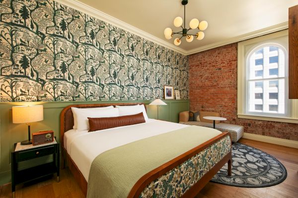 A cozy bedroom with a large bed, decorative wallpaper, modern lighting, and a window with an urban view. The room features a mix of vintage and contemporary decor.