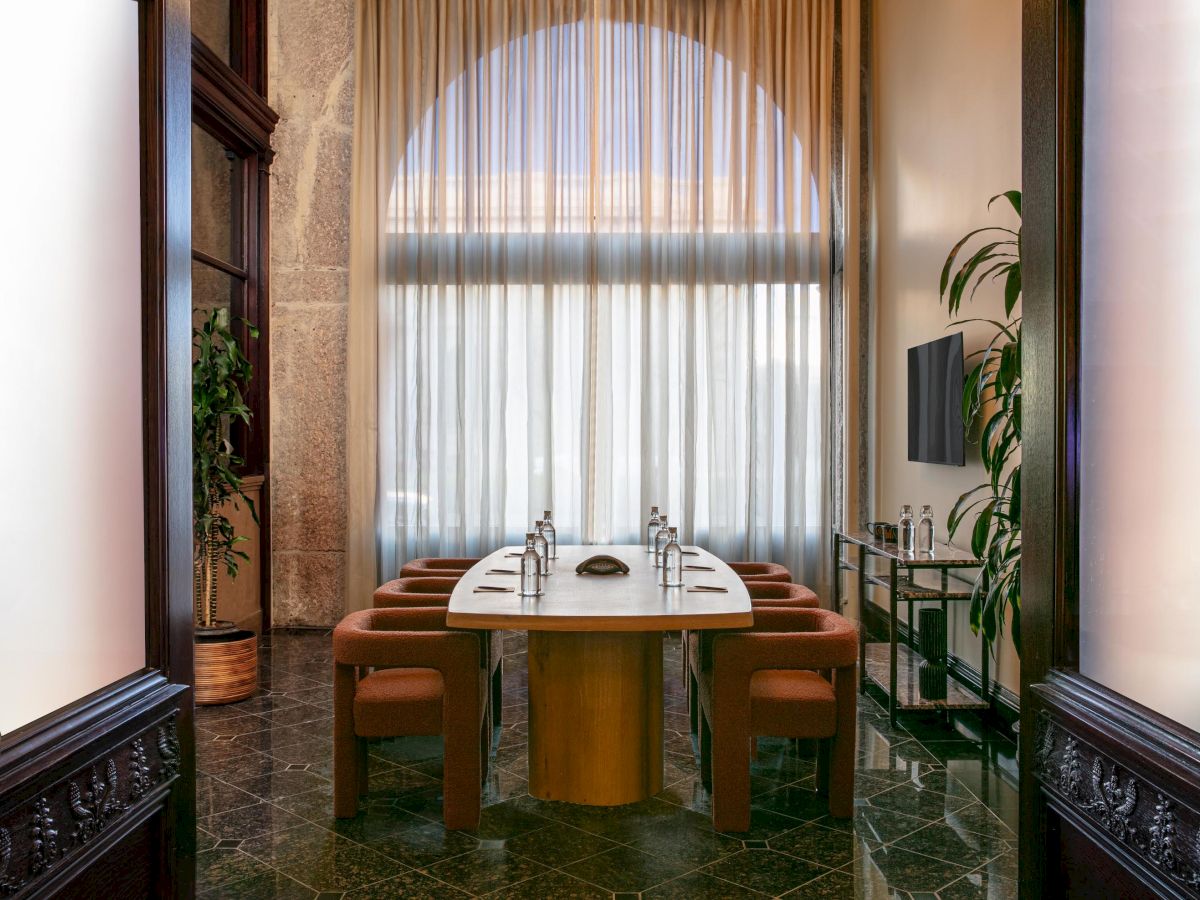 A meeting room with a long table, several chairs, bottled water, plants, and a TV screen, under a large arched window with sheer curtains.