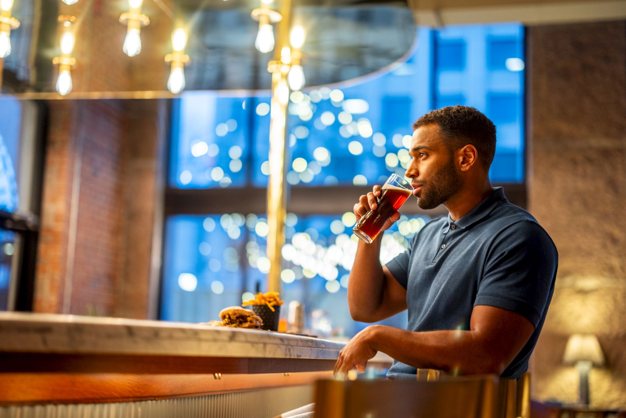 A man in a blue shirt is standing at a bar, holding a drink and looking to his left. There's food on the counter and hanging lights above.