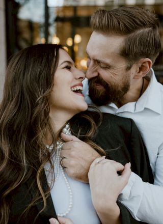 A joyful couple is embracing and smiling at each other, showcasing their happiness and affection in an outdoor setting with blurred lights in the background.