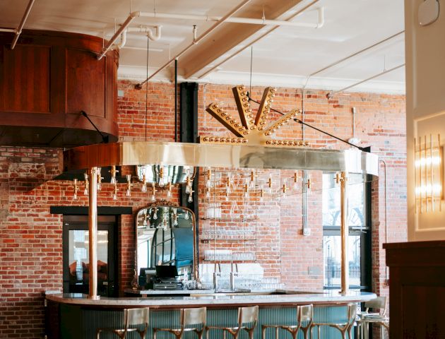 The image shows a cozy bar area with brick walls, a long counter, metal bar stools, hanging lights, and a window with natural light.