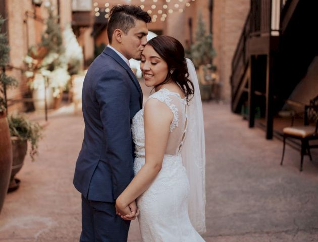 A bride and groom stand close together in an outdoor venue with string lights overhead. They are dressed in wedding attire and holding hands.