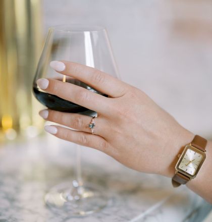 A hand with a wristwatch and ring holding a glass of red wine, resting on a marble surface.