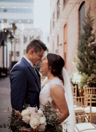 A couple, dressed in wedding attire, stands close together in an outdoor setting, with string lights and chairs in the background, holding a bouquet.