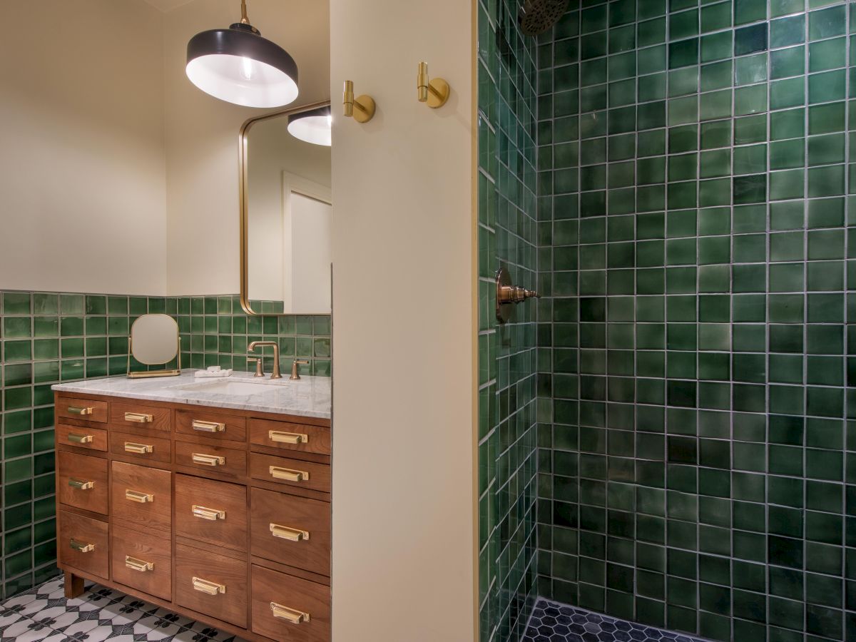 The image shows a bathroom with green tiled shower walls, wooden vanity with brass handles, large mirror, and a black and white tiled floor, ending the sentence.