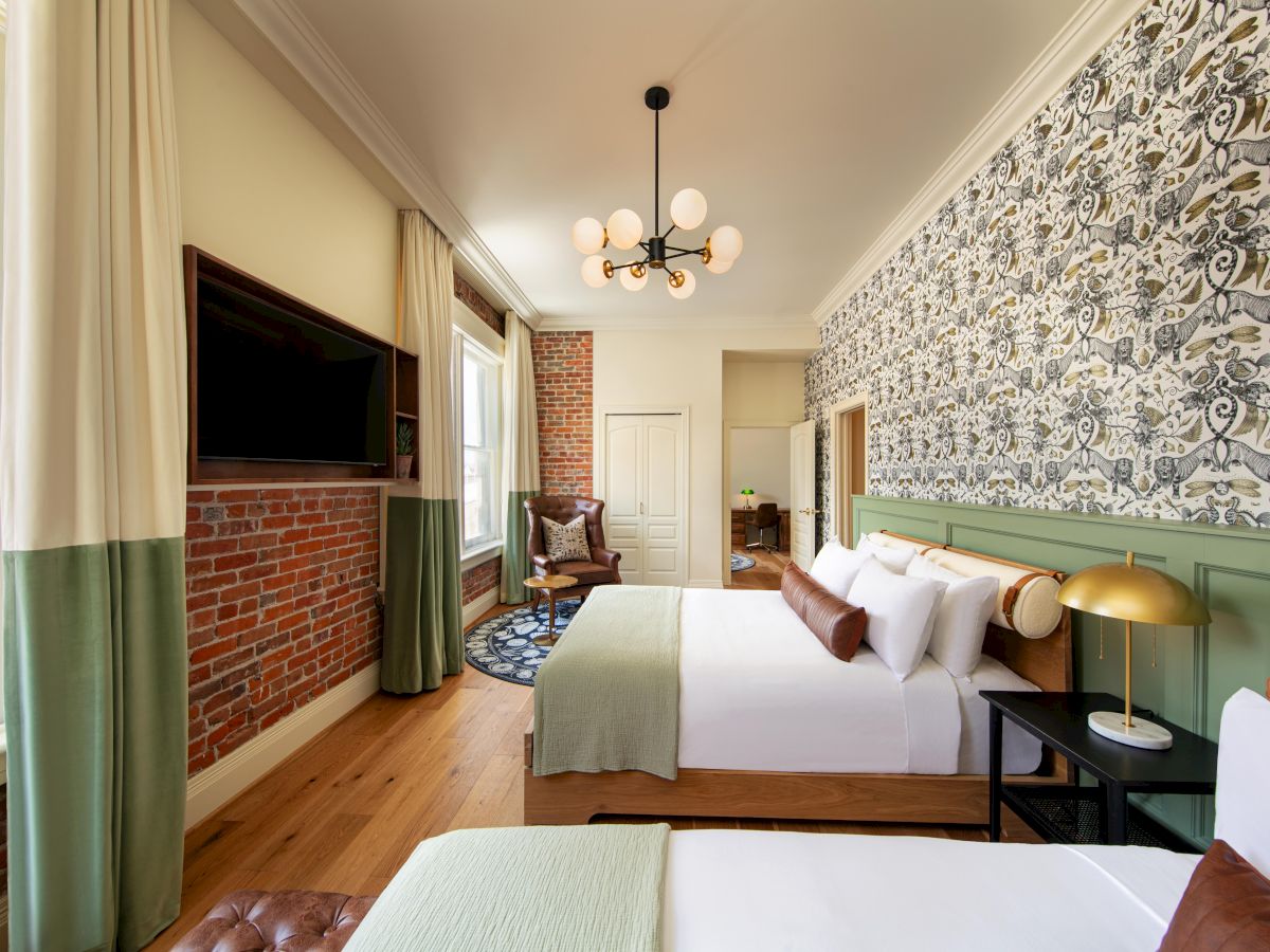 A stylish bedroom with two beds, a large window, a patterned accent wall, a mounted TV, a modern chandelier, and warm decor.