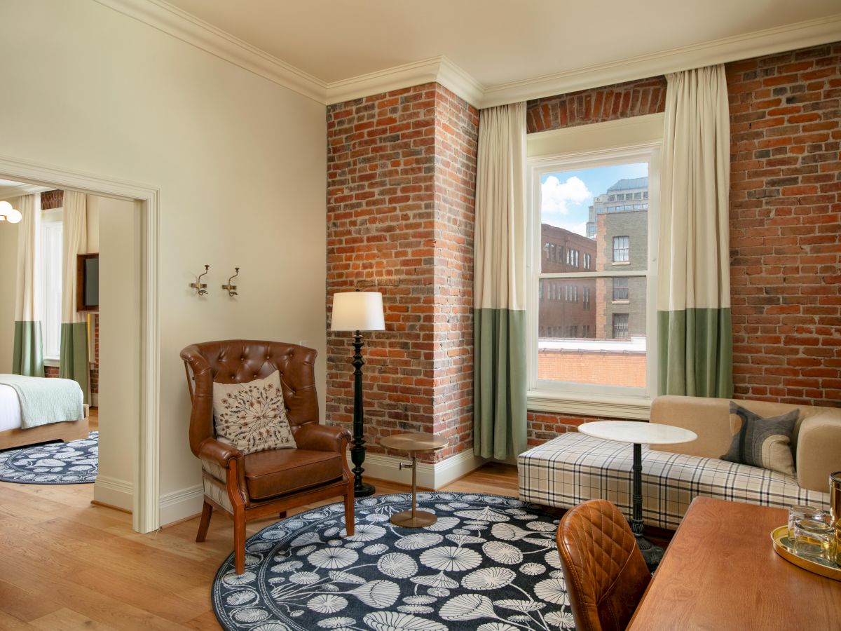 A cozy living area with exposed brick walls, a leather armchair, a round rug, a small table with chairs, and a view of buildings through the window.