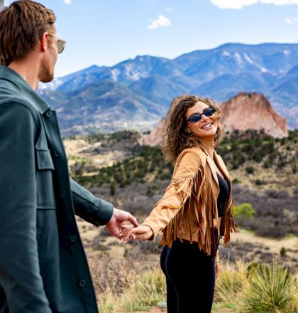 A couple, holding hands, with a scenic mountain backdrop and clear sky. The woman is smiling and wearing sunglasses.