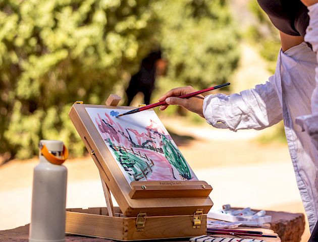 A person is painting a landscape outdoors with an easel, paint, and brushes. Sunglasses and a water bottle are on the table.