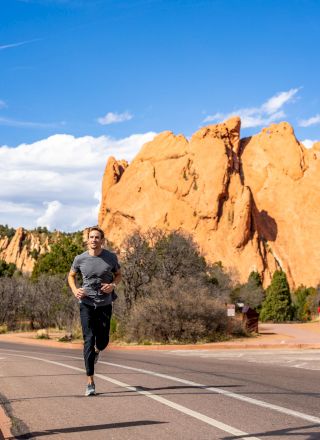 A person is jogging on a road with large rock formations and clear blue skies in the background.