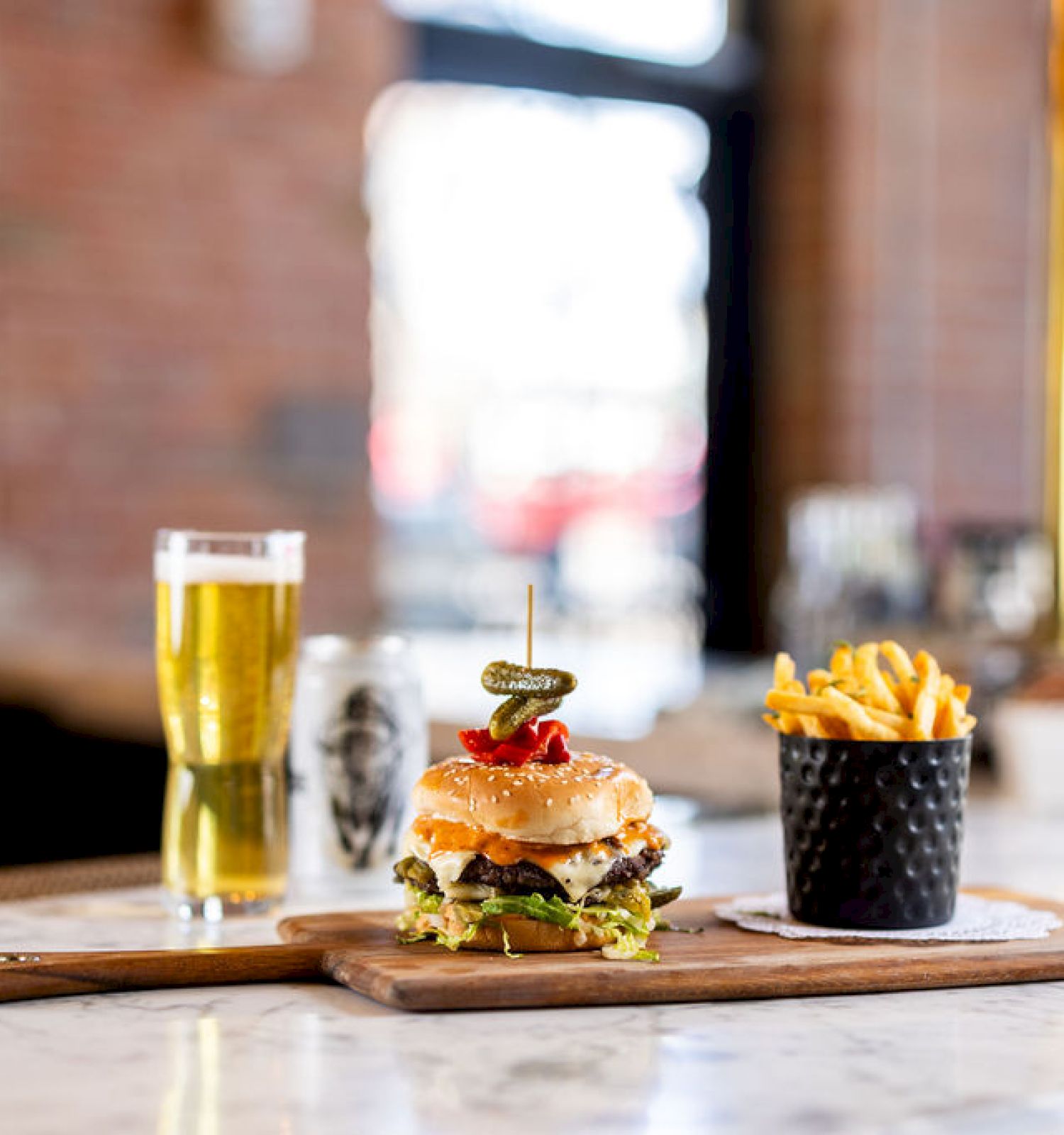 A burger and fries on a wooden board, accompanied by a glass of beer, are placed on a marble countertop in a cozy, indoor setting.